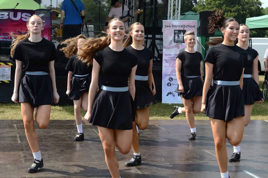 A group of people in black outfits performing on stage, mid irish dance. They are mid-move all having their left foot raised.