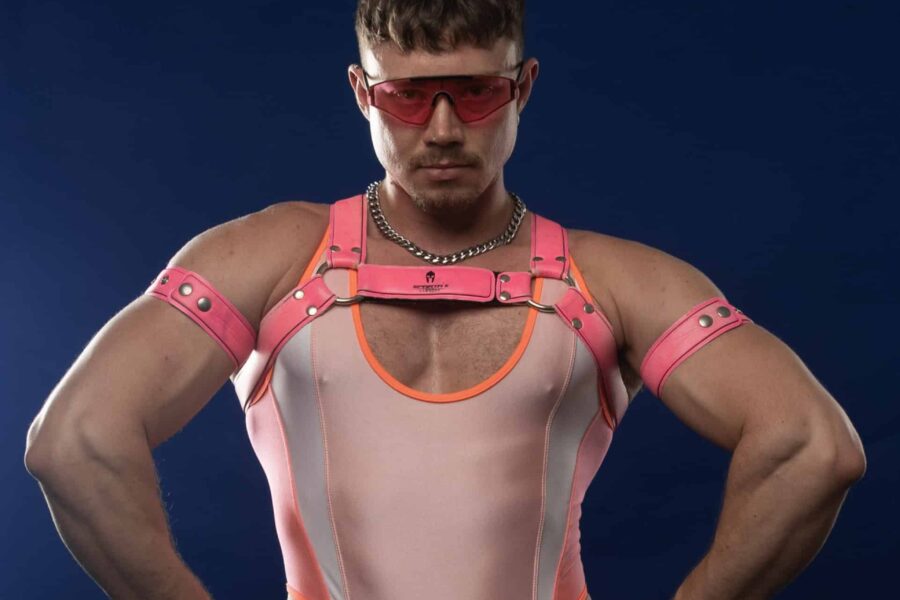 A performer in front of a dark blue backdrop, they are wearing a pink leotard style outfit, with neon pink details. They are posed like a body builder.