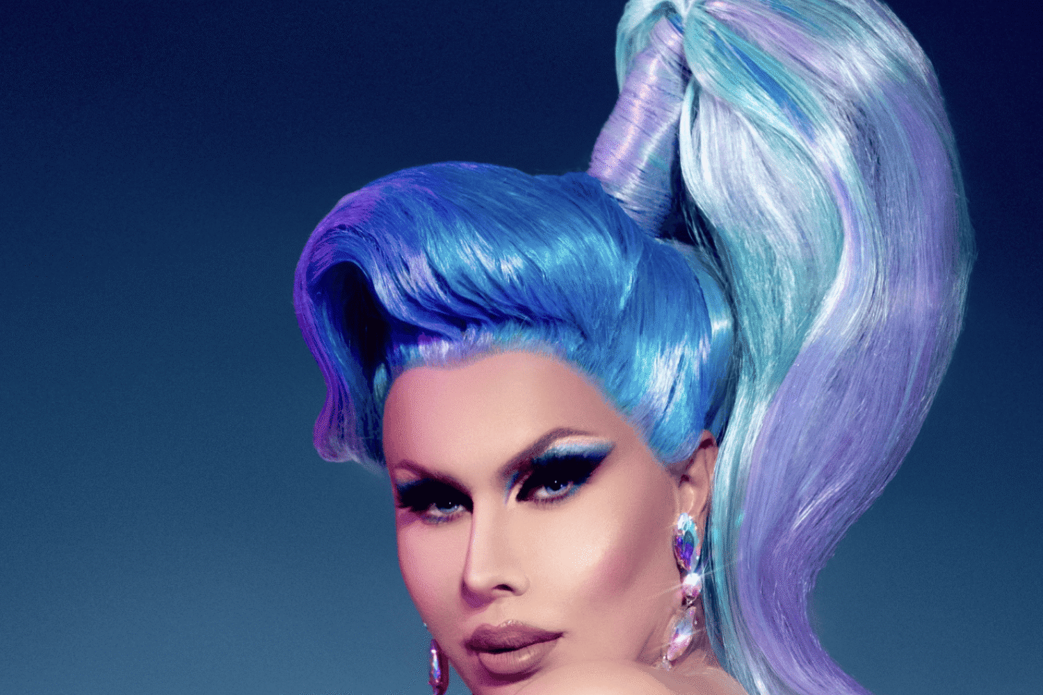 Image if Trinity The Tuck in a blue wig with high pony tails. She is side on but her face is toward the camera and her outfit drapes off one shoulder.