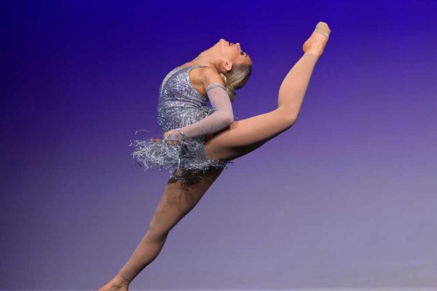 A performer mid-leap on stage. There is a blue to white gradient backdrop, they are mid-move, with their right leg extending, and their left arched up behind them, and their head tilted back.
