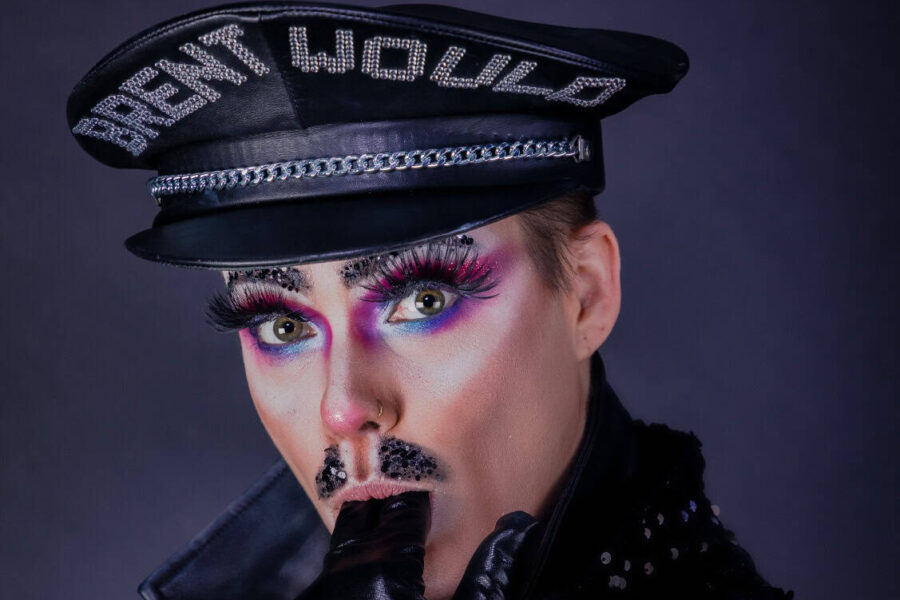 A performer dressed in a leather outfit in a mid-close up photo. They are wearing a police style hat that has 