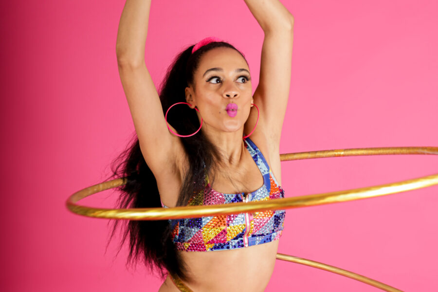 A performer in front of a pink background mid hula hoop routine. The performer has long brown hair tied in a pony tail and is wearing an 80s style athletic outfit.