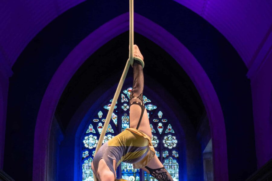 A performer in a church, they are suspended from an aerial rope. They have long blonde hair.