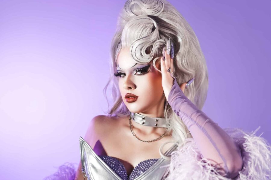 Darcii Dalore on a purple background wearing a silver corset with feather frinding and a blonde wig. She has one had up to the side of her head