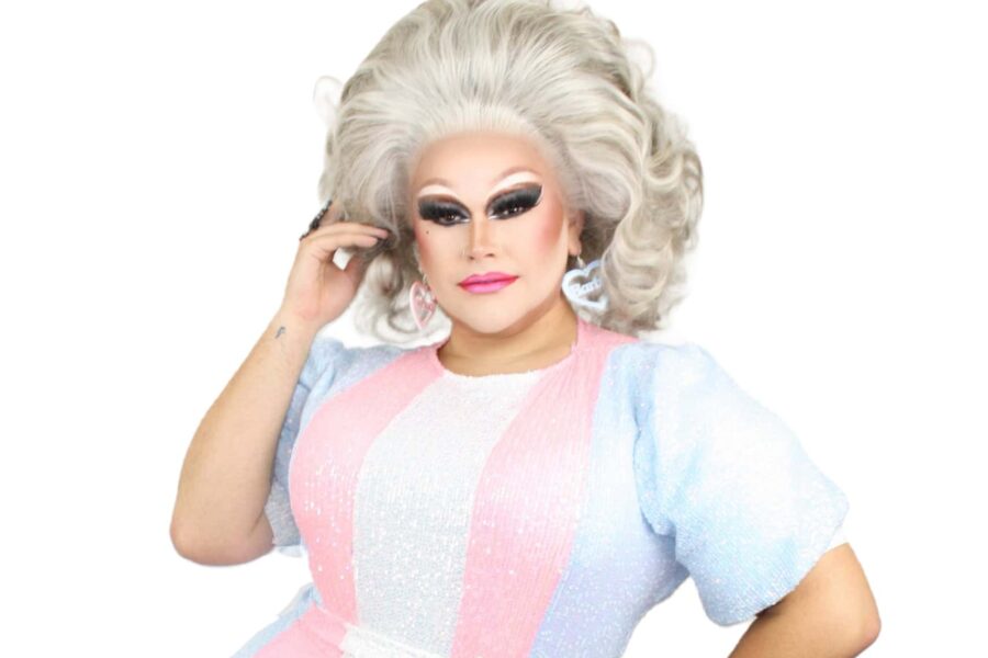 Dixie stands on a white background and wearing a sequin doll style dress in the trans colours and a blond wig.