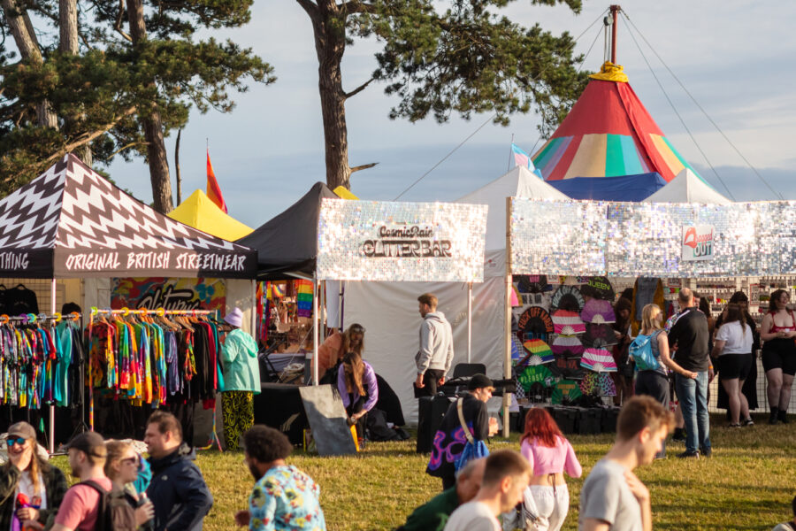 Images of traders trader in the distance with people in the foreground at the festival. Behind the stalls you can see two trees and the top of a rainbow marquee.