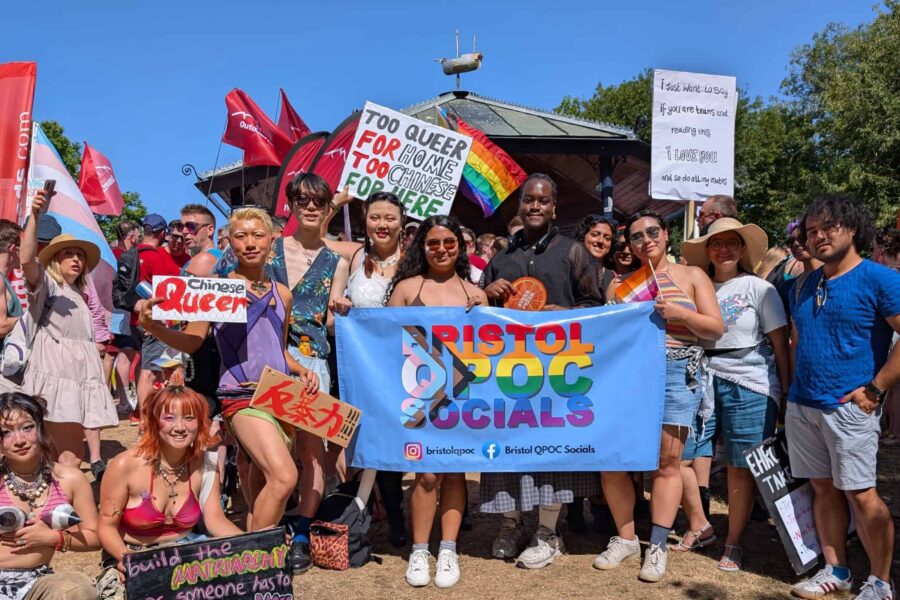 A group of people stood outdoors in the sun smilikng at the camera, there is a large banner held up in the centre of the people that reads Bristol QPOC socials.