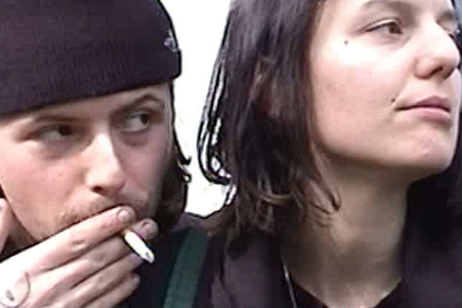 An image with two people, the person on the left is wearing a beanie and smoking a cigarette, the person on the right is looking off to their left and has medium length straight brown hair.