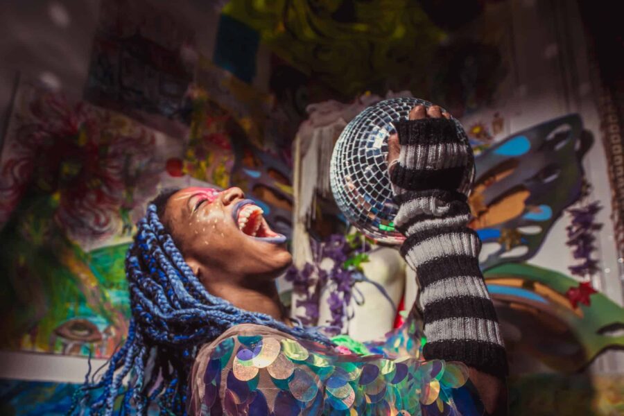 An eclectic scene with Juice NazNaz as the focus, with long blue briads, a scaly iridescent jacket and a huge grin on their face as their head tips back, holding a mirrorball almost like a mircophone.