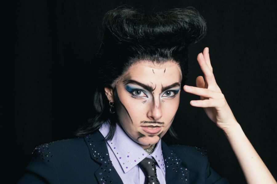 A pompodoured artist staring at the camera with makeup that makes them look almost like a cartoon, with tiny pen streaks highlighting their features. They are wearing a suit with tiny scattered diamonds, and posing their hand to their face like a pop star in a photo shoot.