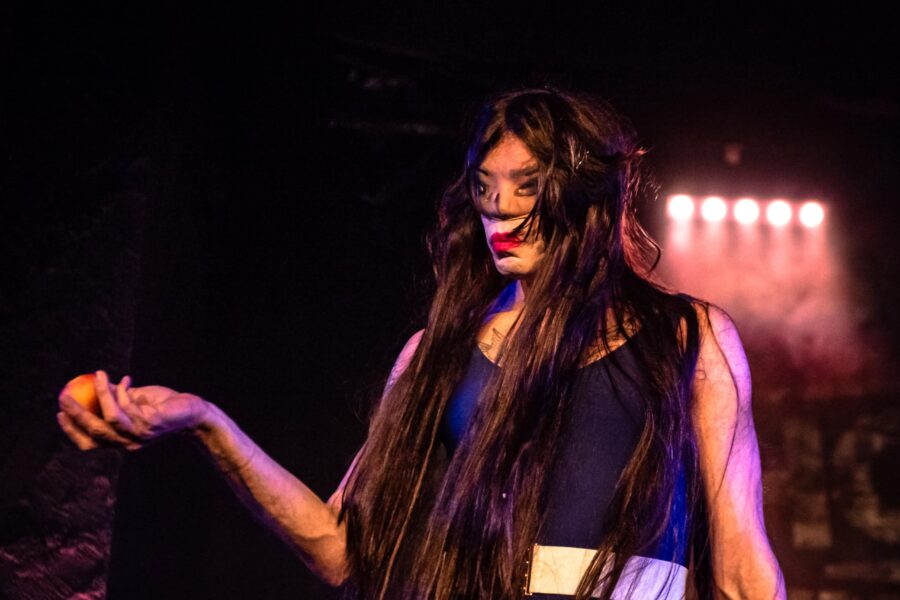 A long haired artist in a purple 80s style leotard holding an apple in their outstretched arm. Their face is made up and they stare out at the audience with a indesiphrable emotion.